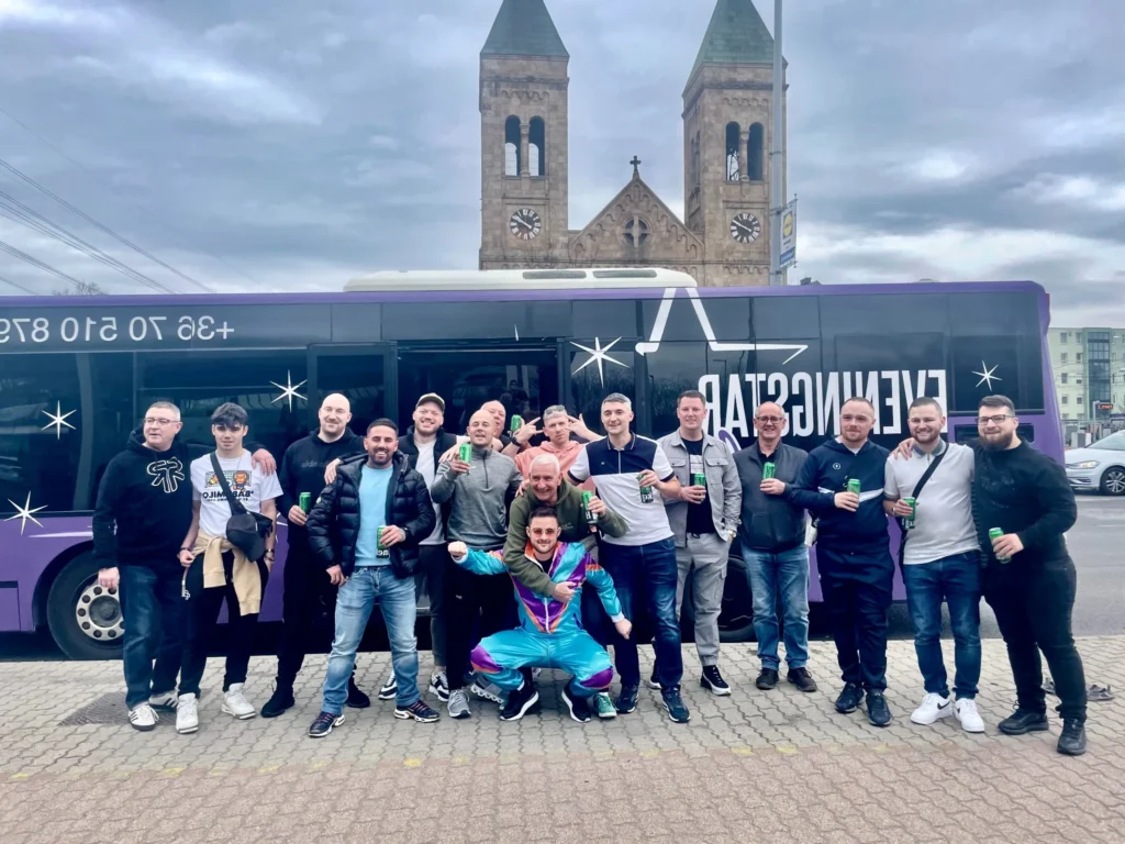 Group of friends posing in front of a purple party bus, holding drinks during a daytime pickup in Budapest.