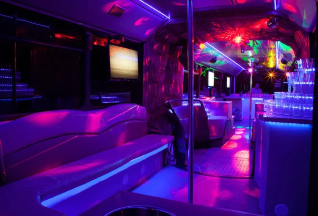Party bus interior with leather lounge seating, neon LED lighting, and a bar area with glassware.